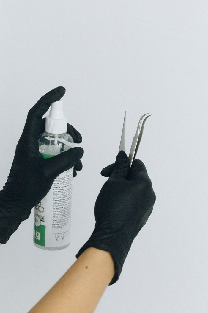 Black gloved hands holding a medical spray bottle and tweezers on a white background.